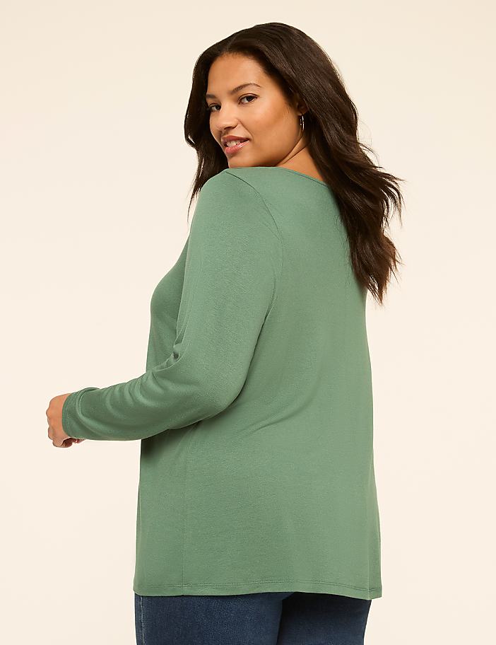 A woman in a green long-sleeved shirt and blue jeans poses against a plain, light-colored background.