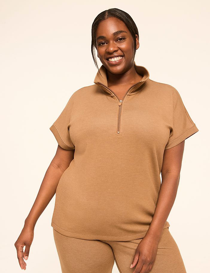 A smiling woman in a brown quarter-zip top and matching pants stands against a plain, light-colored background.