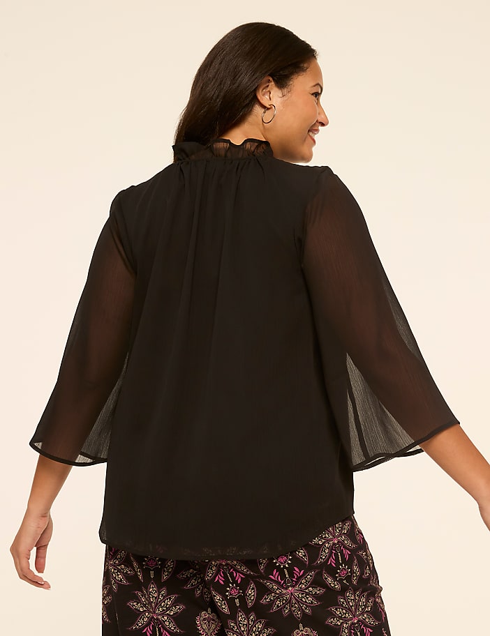 A woman in a black ruffled-neck blouse and patterned skirt smiles over her shoulder against a plain background.