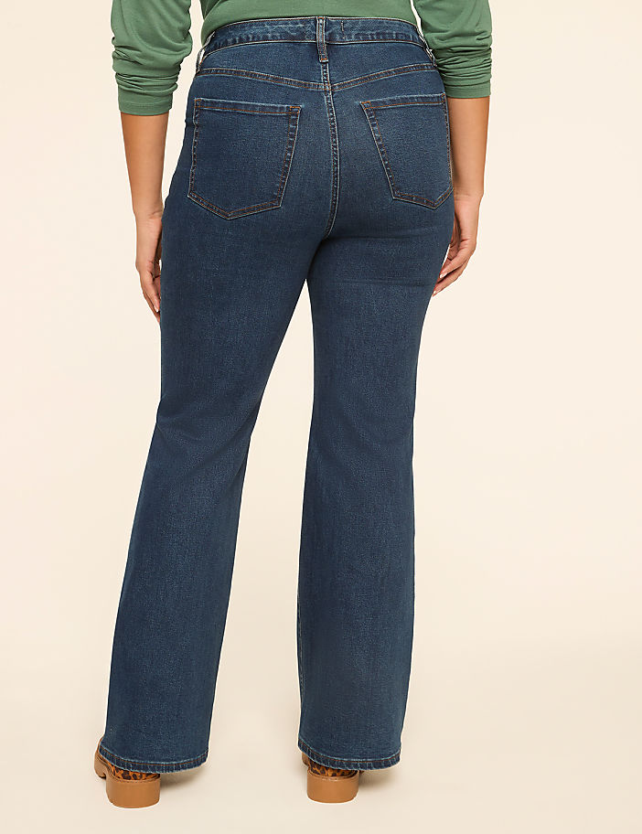A person wearing dark wash flare jeans stands against a plain, light-colored background.