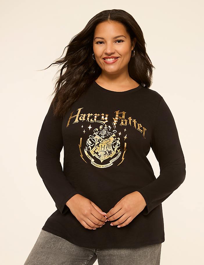 A smiling woman with dark, wavy hair wears a black Harry Potter t-shirt with a gold crest and text, standing against a plain, light-colored background.