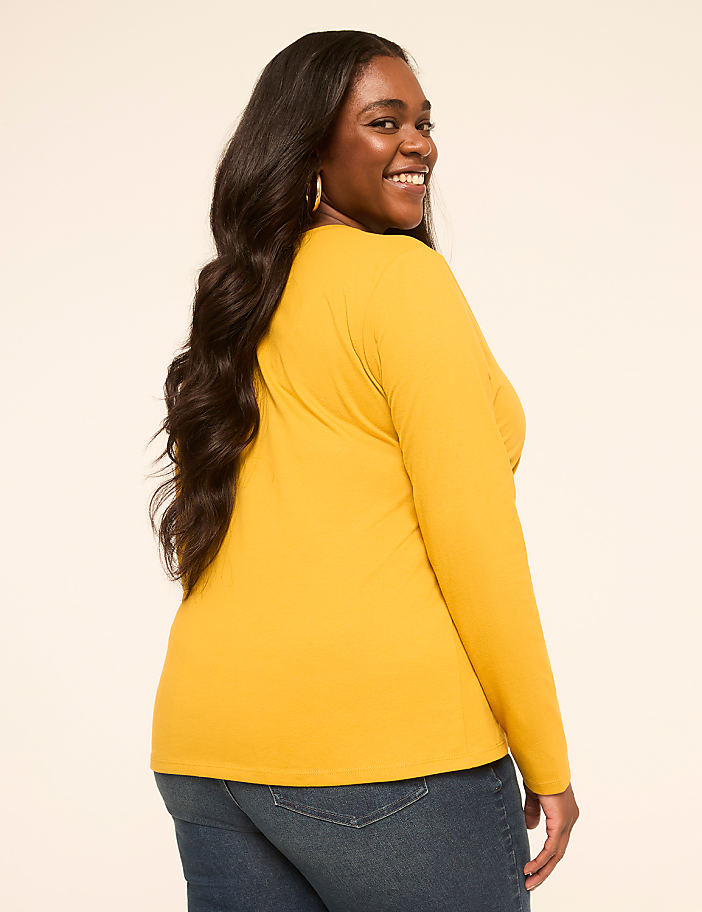 A smiling woman with long dark hair, wearing a mustard yellow long-sleeved shirt and blue jeans, is shown from the side against a plain, light-colored background.