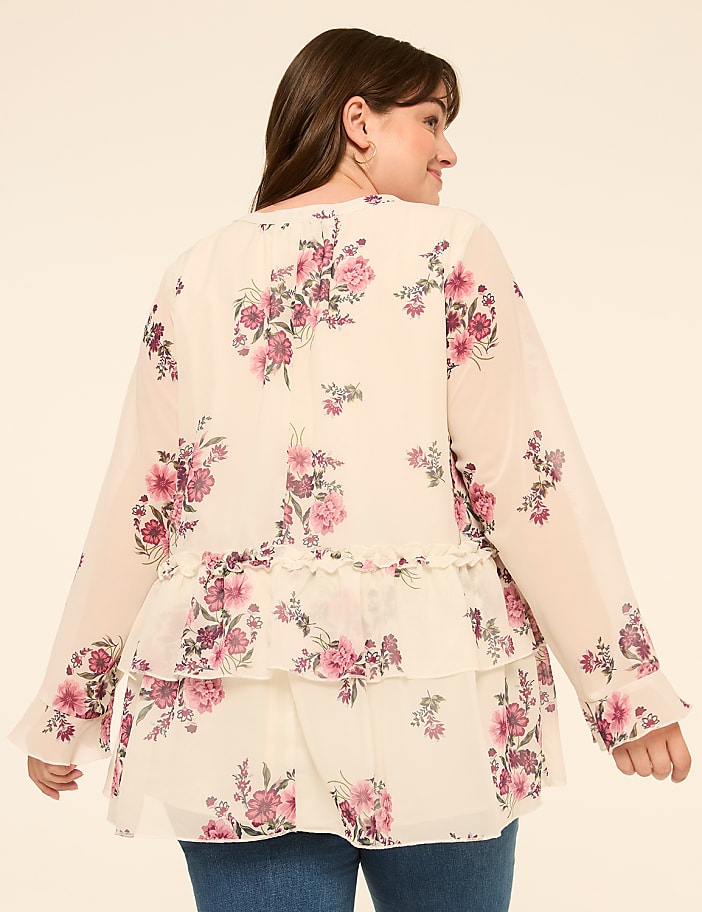 A woman in a floral print blouse with ruffled tiers stands against a plain, light-colored background.