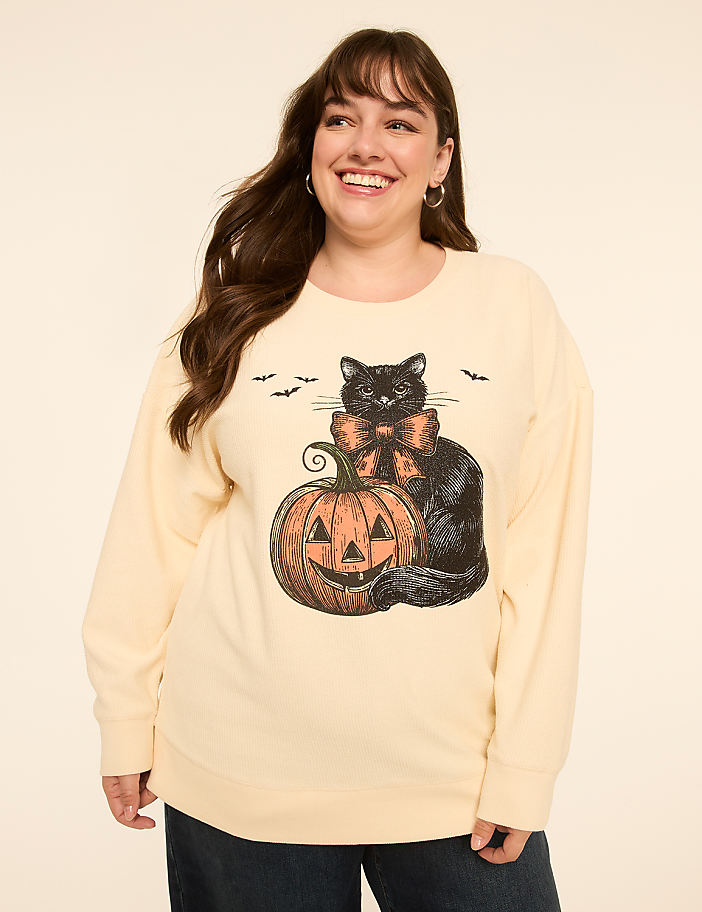 A smiling woman in a cream-colored sweatshirt with a Halloween graphic of a black cat and pumpkin stands against a plain, light background.
