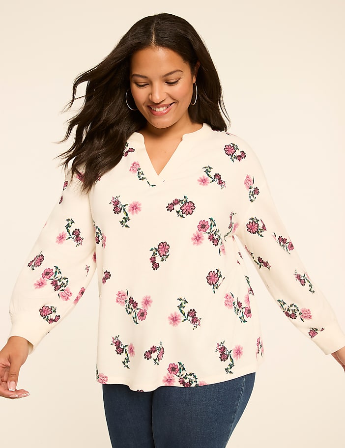 A smiling woman in a cream-colored floral sweater and blue jeans stands against a plain, light background.