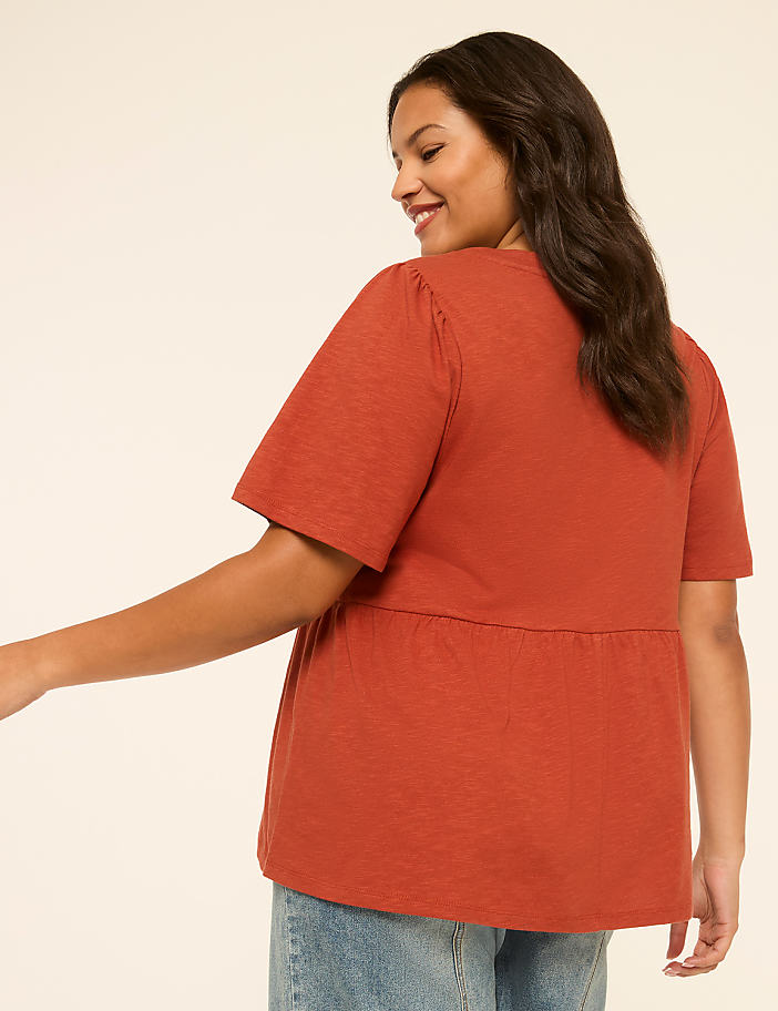 A woman in a rust-colored peplum top and jeans smiles over her shoulder against a plain, light background.