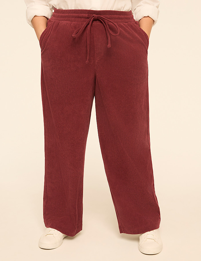A person stands with hands in the pockets of maroon corduroy pants, against a plain, light-colored background.