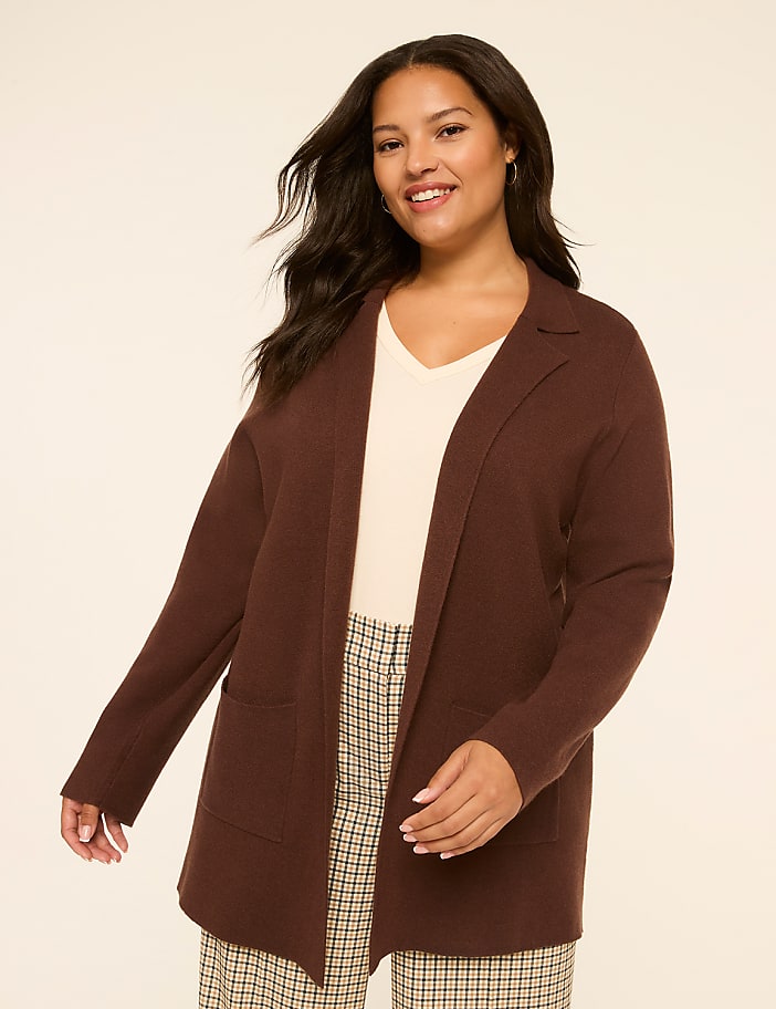 A smiling woman in a brown cardigan and plaid pants stands against a plain, light-colored background.