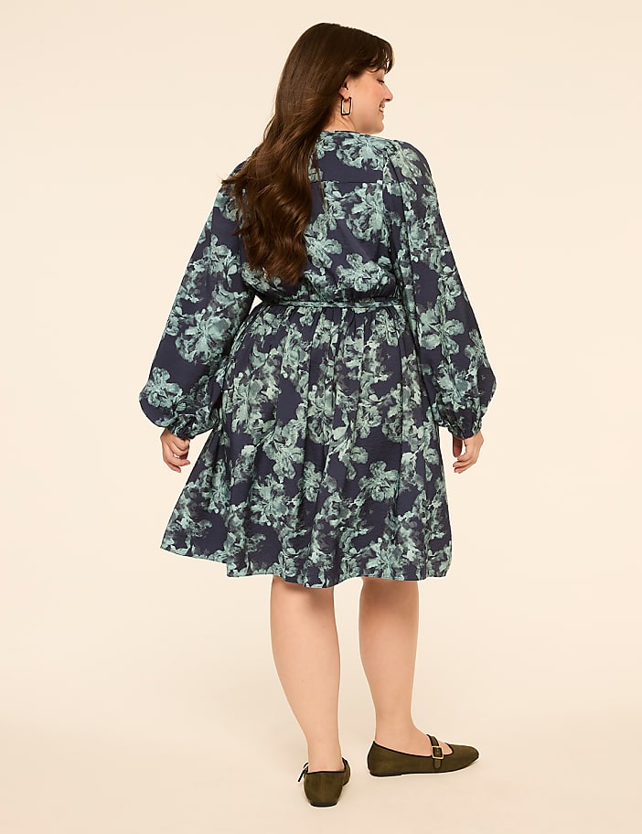 A woman in a floral dress stands against a plain, light-colored background.