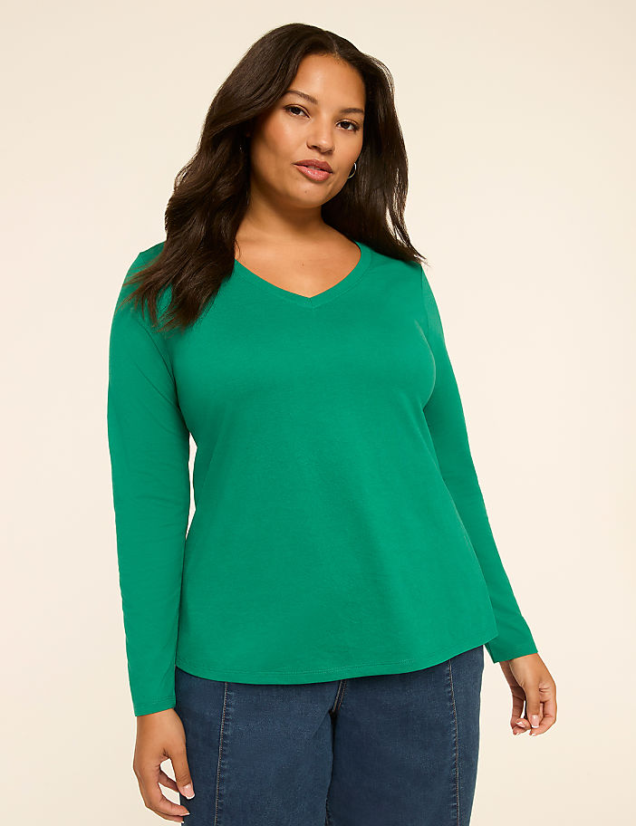 A woman with dark hair and a v-neck green long-sleeved shirt stands against a plain, light-colored background.