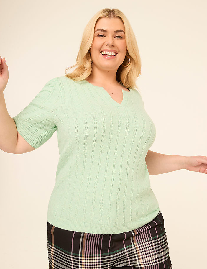A smiling woman in a mint green top and plaid pants poses against a plain, light background.