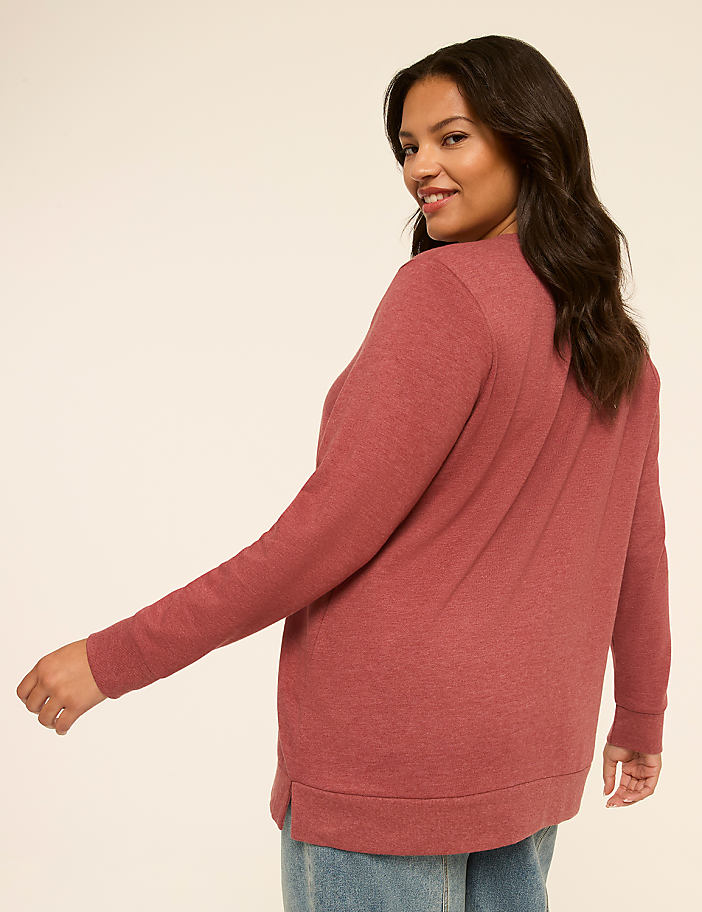 A woman in a rose-colored sweater and jeans turns to smile over her shoulder against a plain, light background.