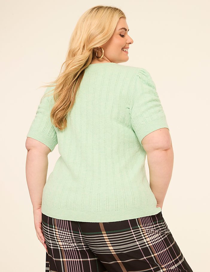 A woman in a light green cable-knit top and plaid pants stands against a plain, light-colored background.