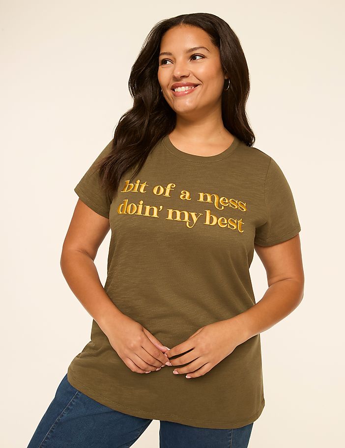 A smiling woman in a green t-shirt with gold lettering stands against a plain, light-colored background.