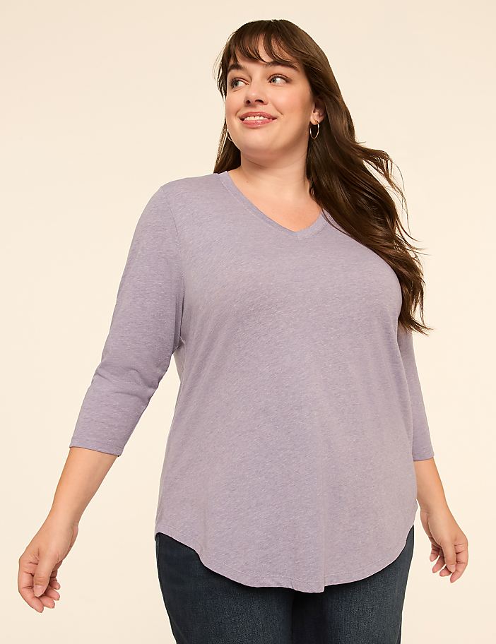 A woman in a lavender v-neck shirt and dark jeans smiles against a plain beige background.