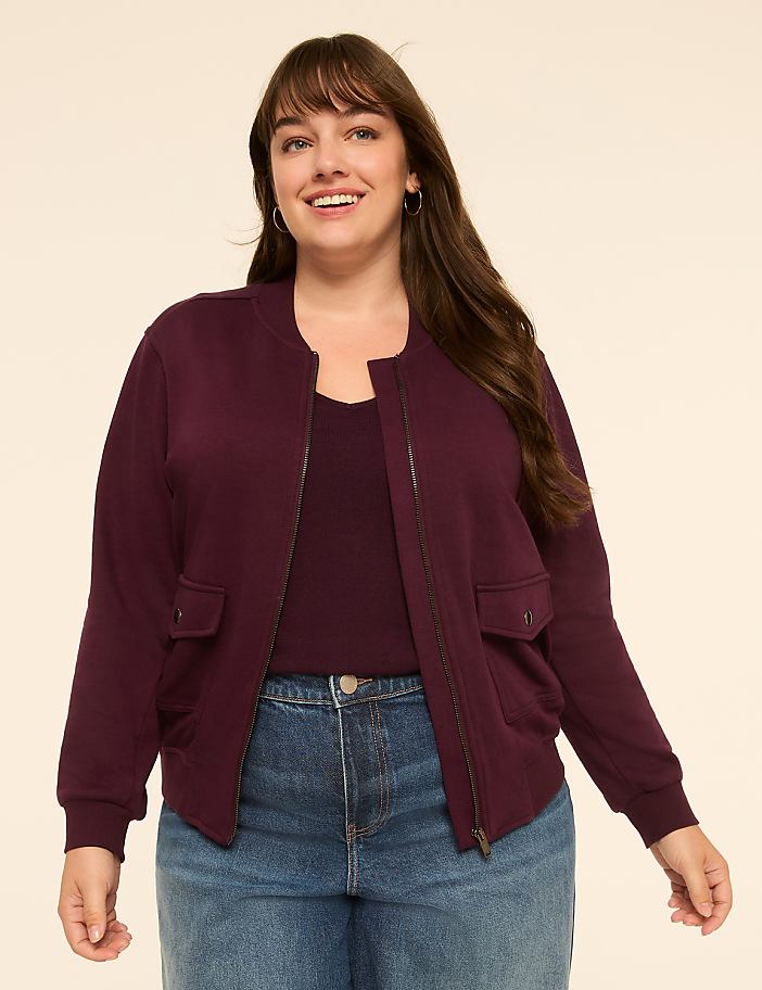 A smiling woman in a burgundy bomber jacket and blue jeans stands against a plain, light-colored background.