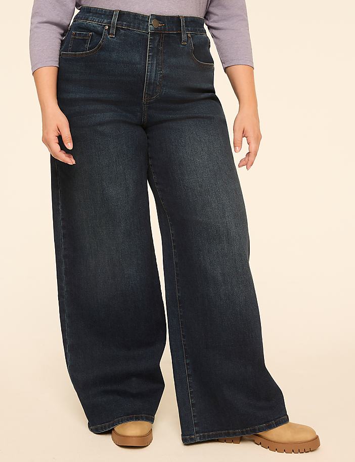 A person wearing dark wash wide-leg jeans and a light purple top stands against a plain beige background.