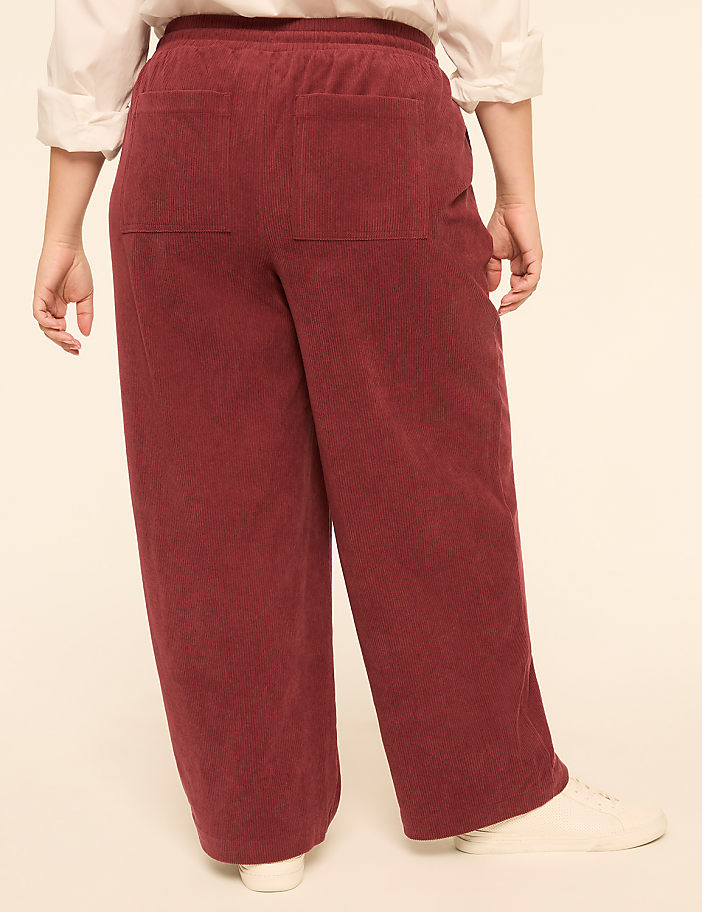 A person wearing maroon corduroy pants and white sneakers stands against a plain, light-colored background.