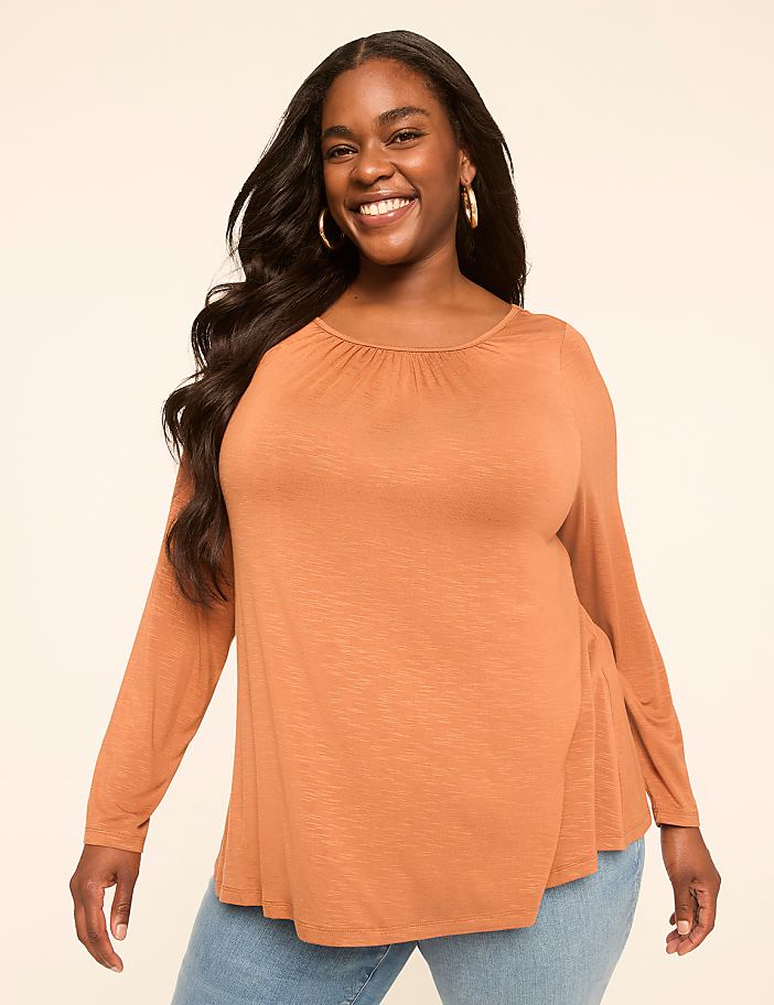 A smiling woman with long dark hair wears an orange long-sleeved shirt and blue jeans against a plain beige background.
