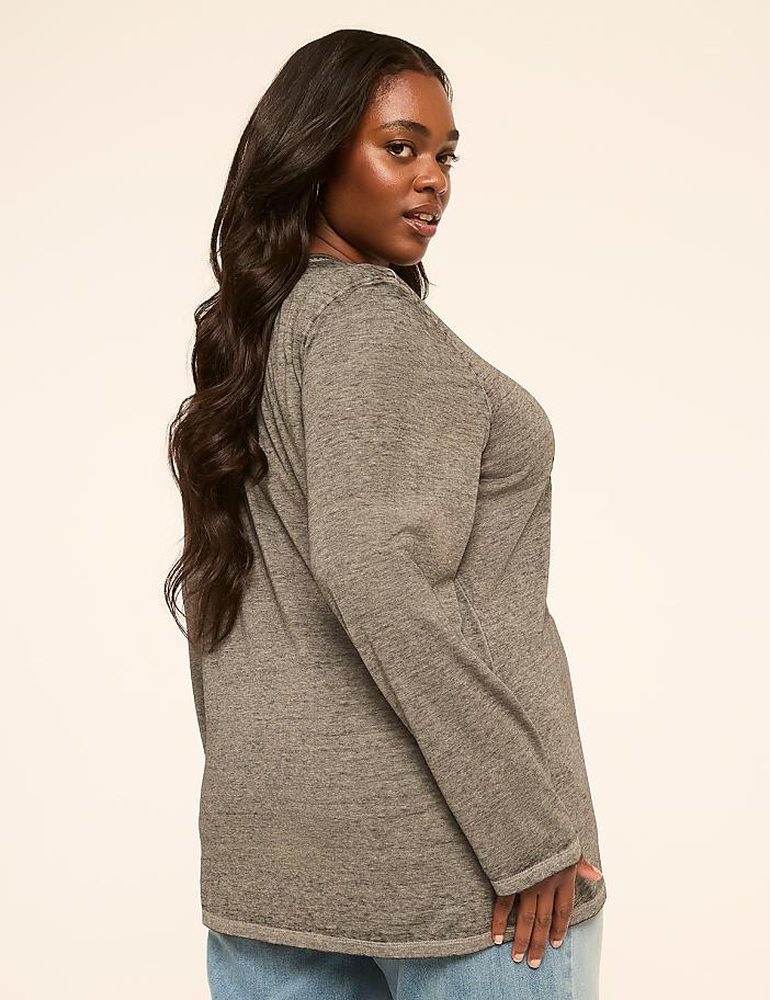 A woman with long, dark hair wearing a gray long-sleeved shirt and blue jeans poses against a plain, light-colored background.