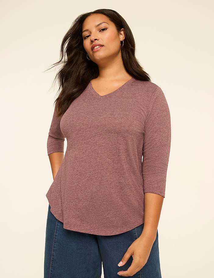 A woman in a mauve v-neck shirt and blue jeans stands against a plain, light peach background.