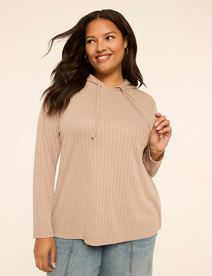 A smiling woman in a beige hooded sweater and jeans stands against a plain, light-colored background.