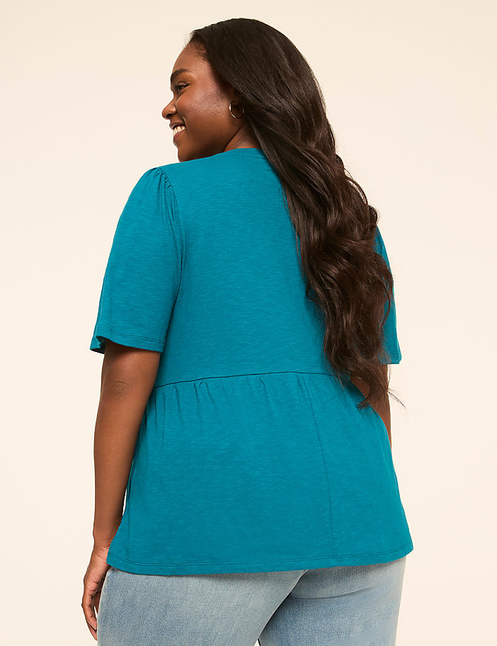 A woman in a teal top and jeans smiles over her shoulder against a plain, light-colored background.
