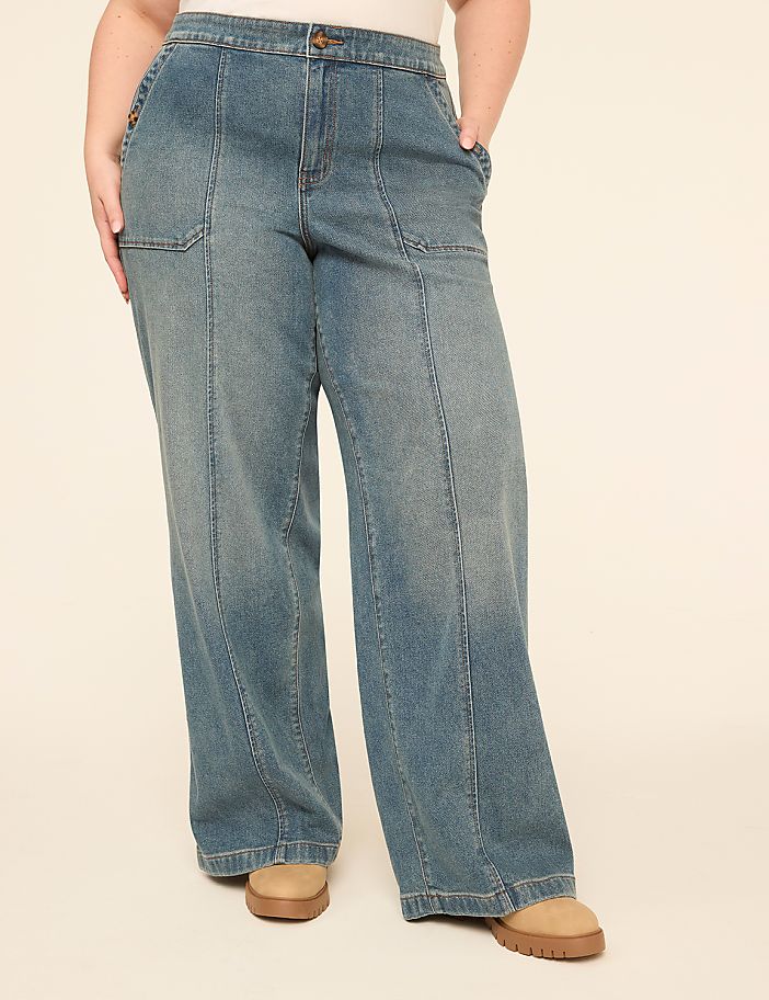 A person wearing wide-leg denim jeans and tan shoes stands against a plain, light-colored background.
