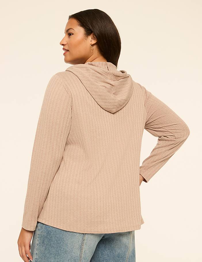 A woman in a beige hooded sweatshirt and jeans stands against a plain, light-colored background.