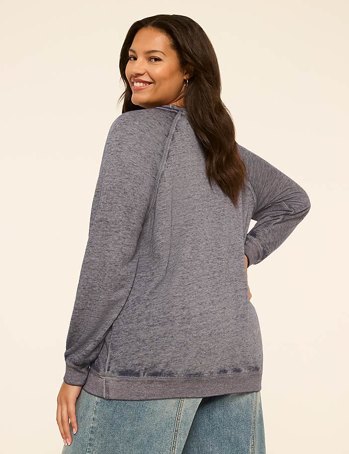 A woman in a blue sweatshirt and jeans stands against a plain cream background.