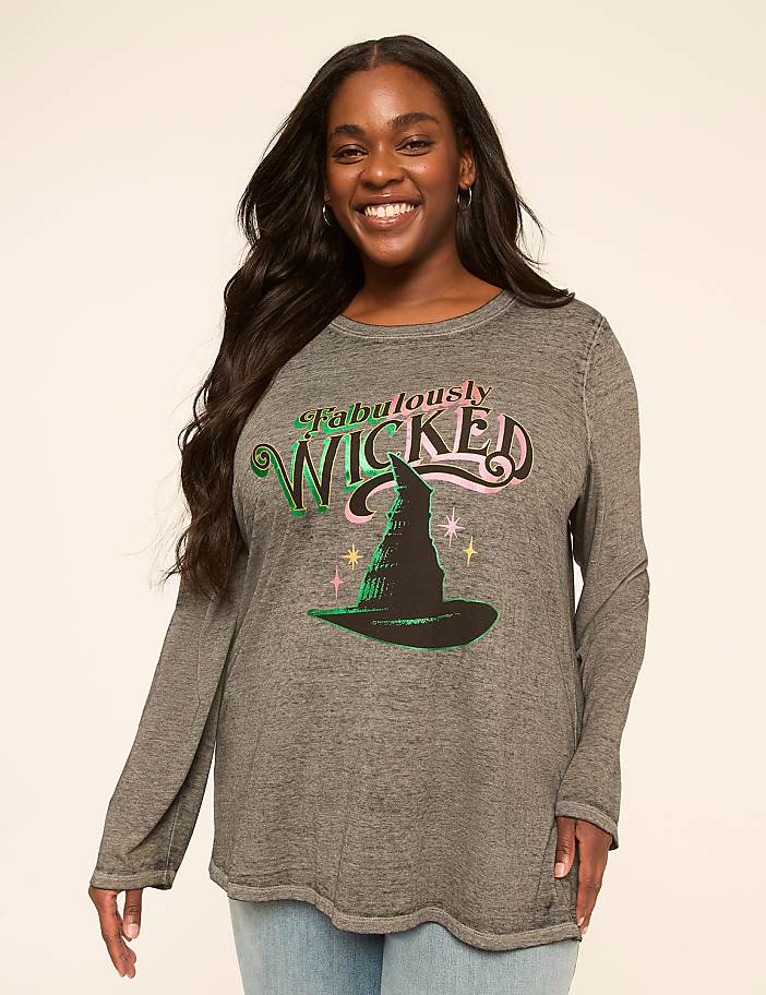 A smiling woman in a long-sleeved shirt with a witch hat graphic stands against a plain, light-colored background.