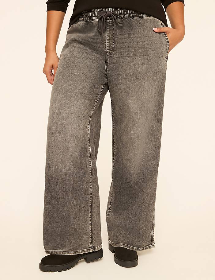 A person stands in wide-leg grey denim pants with a drawstring waist and black boots, against a plain cream background.