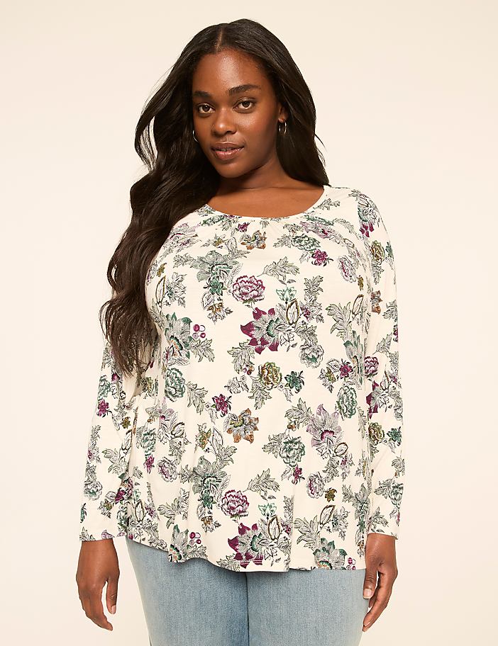 A woman with long, dark hair stands in the foreground wearing a cream-colored, long-sleeved shirt with a floral pattern, against a plain, light-colored background.