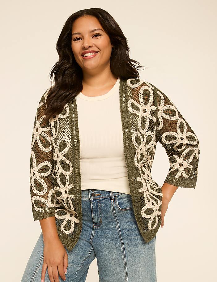 A smiling woman in a floral cardigan and jeans stands against a plain, light-colored background.
