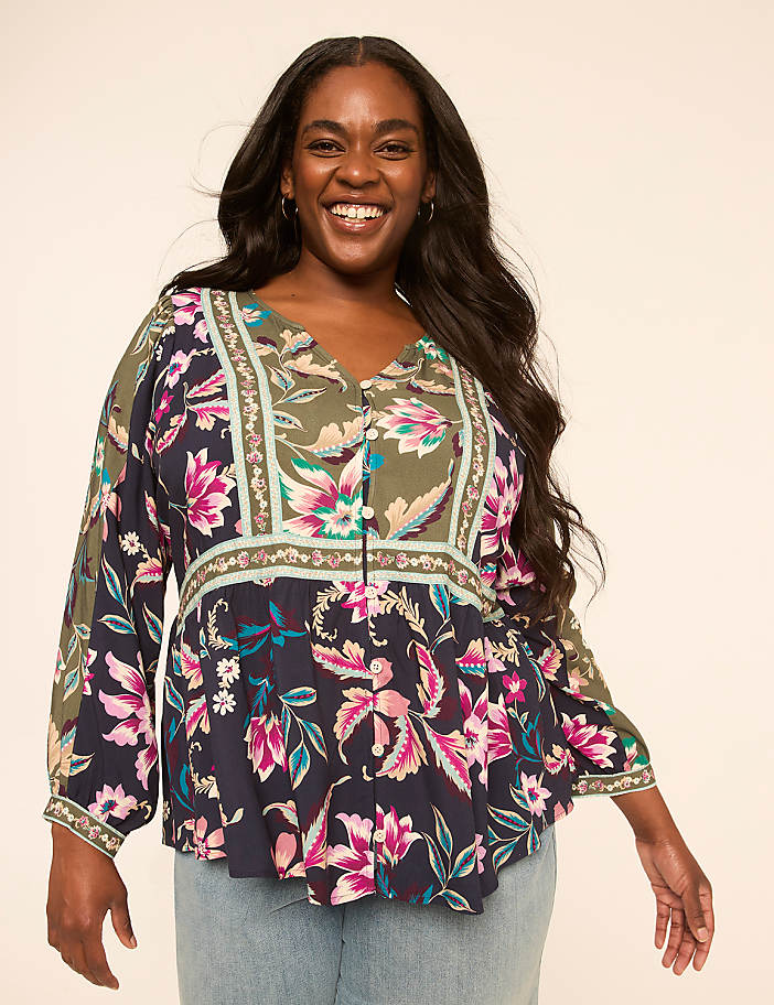 A smiling woman with long dark hair wears a colorful floral blouse and light blue jeans against a plain, light background.