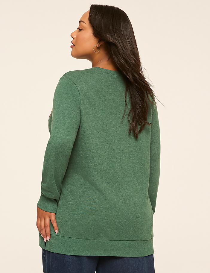 A woman in a green sweater stands against a plain, light-colored background.