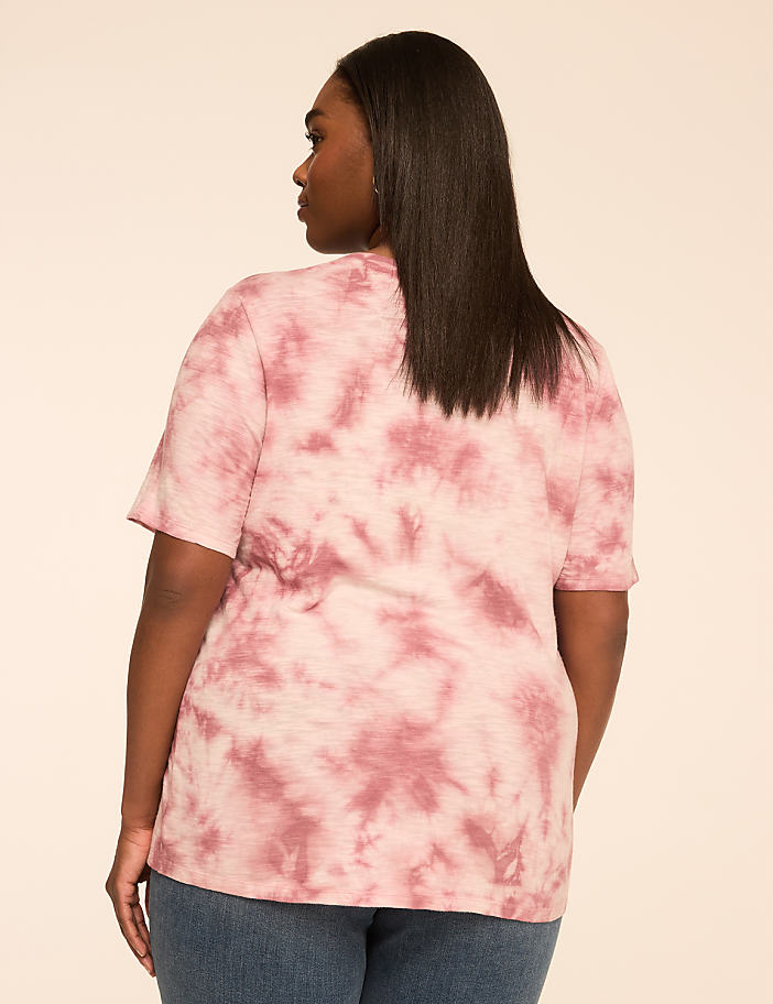 A woman in a pink tie-dye t-shirt and blue jeans stands against a plain, light brown background.