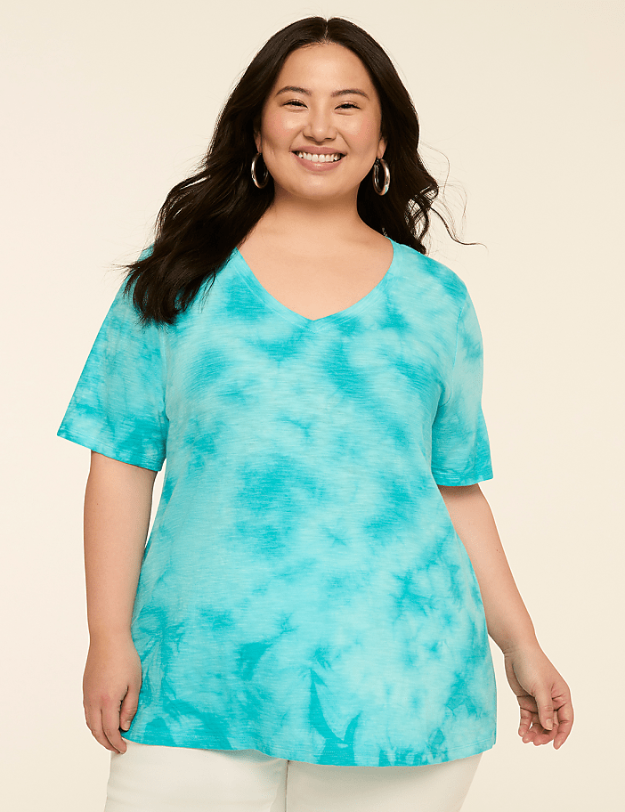 A smiling woman in a turquoise tie-dye v-neck shirt stands against a plain, light-colored background.