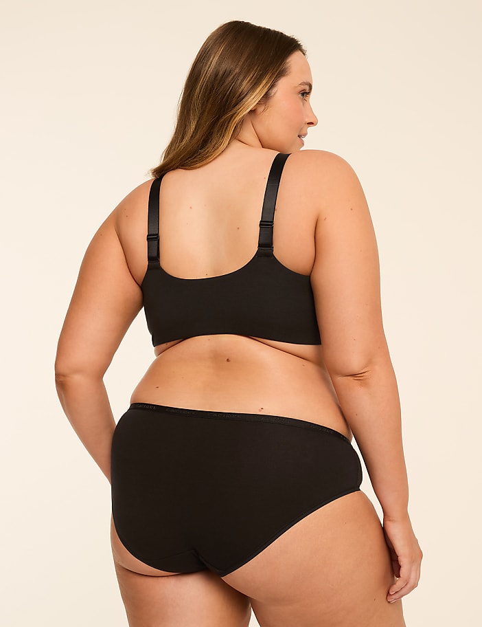 A woman in black lingerie stands against a plain, light-colored background.