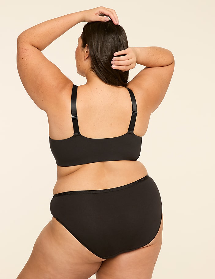 A woman in black lingerie stands with her back to the viewer, her arms raised behind her head, against a plain, light-colored background.