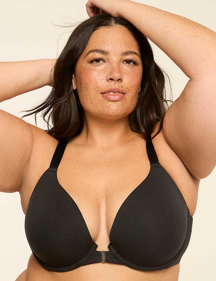 A woman with dark hair and freckles poses in a black bra against a plain, light background.