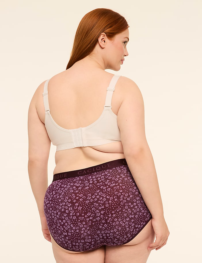 A woman with red hair stands facing away from the camera, wearing a beige bra and purple floral underwear against a plain, light background.