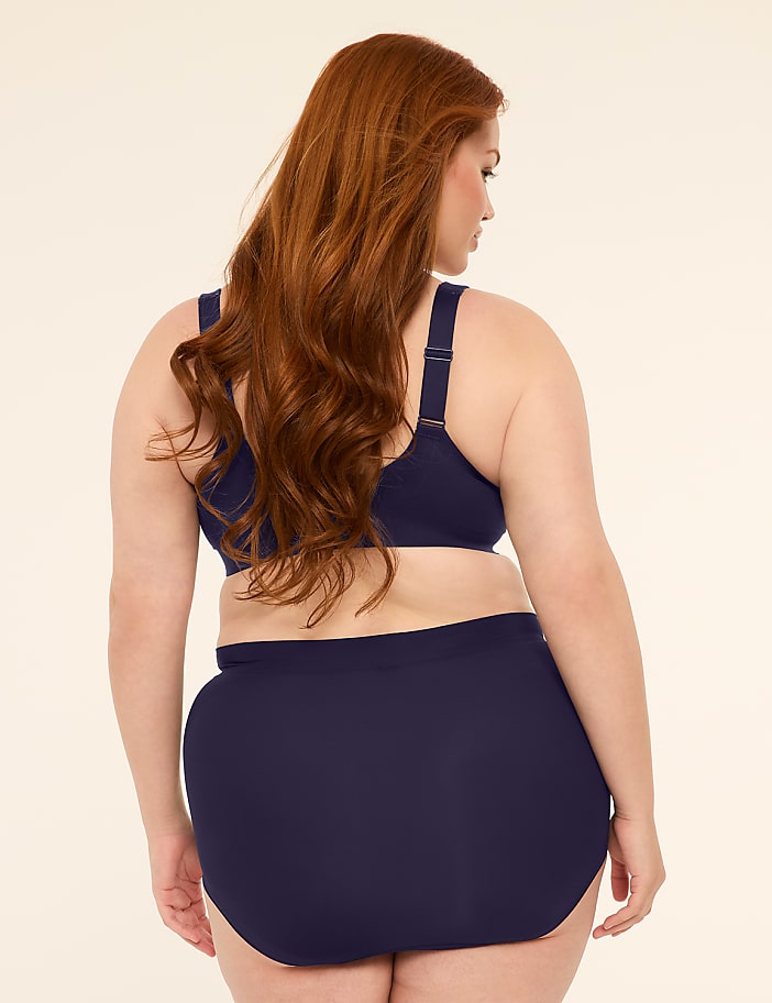 A woman with long red hair stands facing away from the camera in a navy blue bikini against a plain, light-colored background.