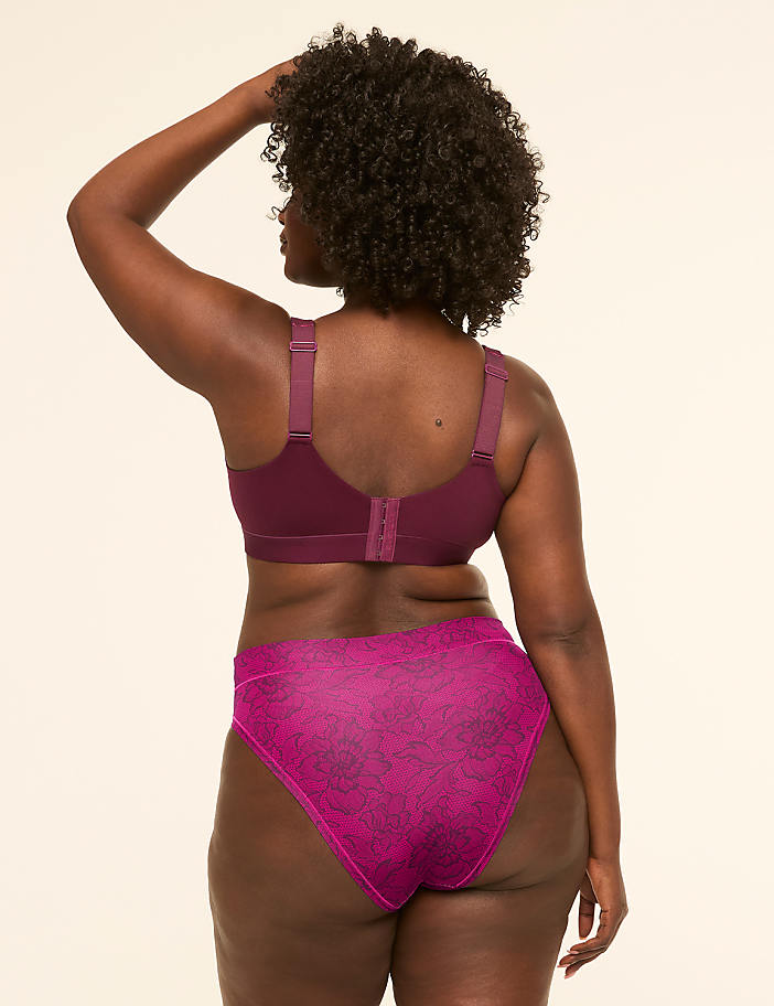 A woman in a burgundy bra and patterned pink underwear stands against a plain, light-colored background.