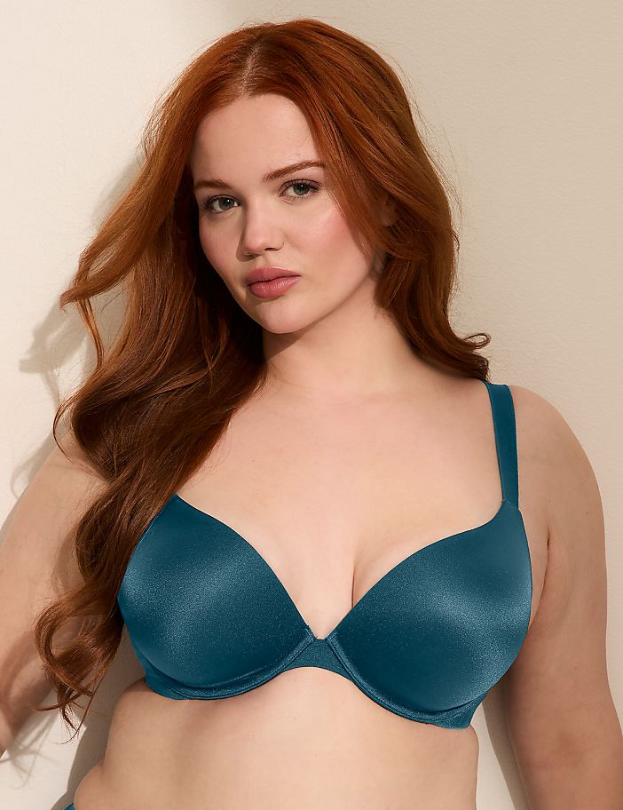 A woman with long, wavy red hair poses in a teal bra against a plain, light-colored background.