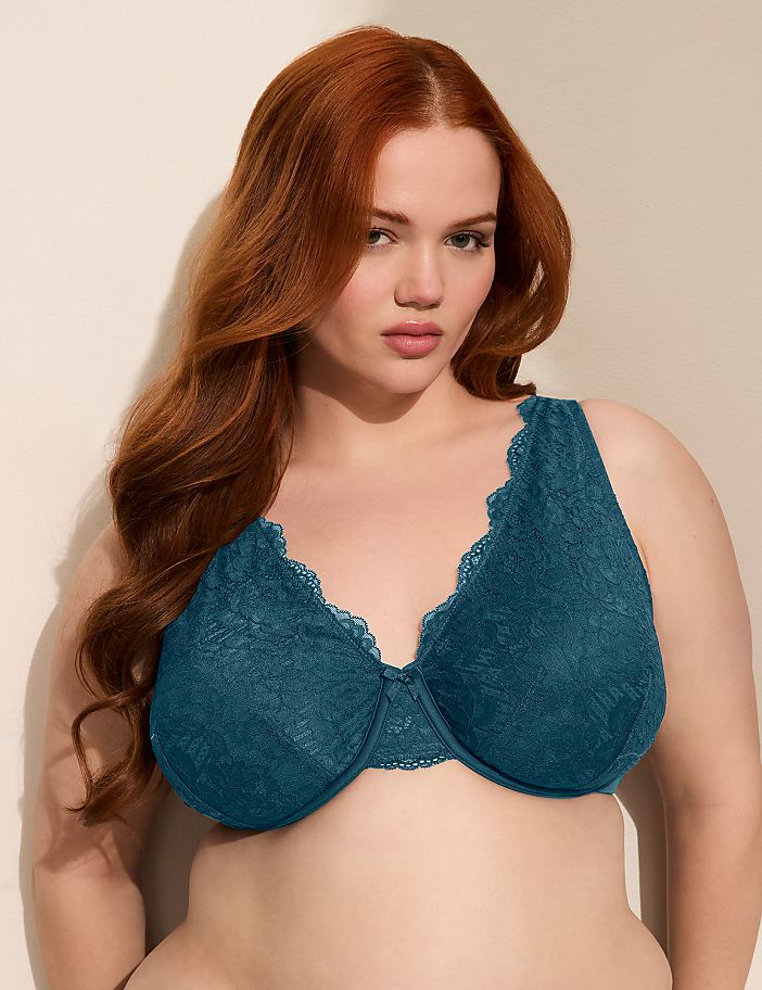A woman with long, wavy red hair poses in a teal lace bra against a plain, light-colored wall.