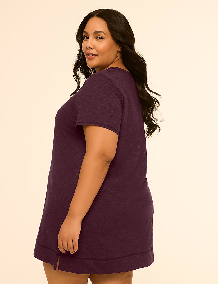 A woman in a purple t-shirt dress stands against a plain, light-colored background.