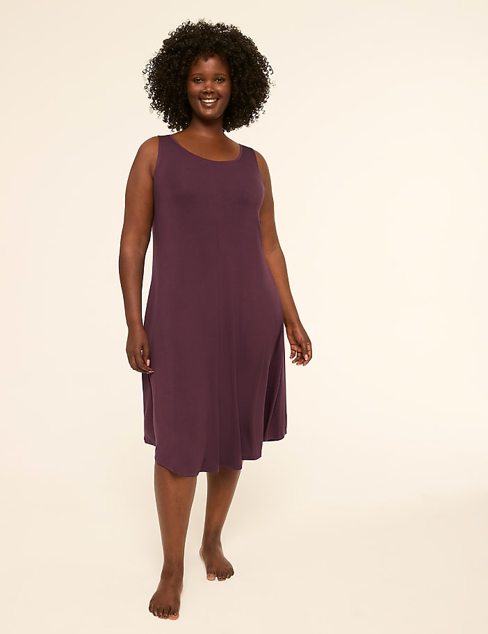 A smiling woman in a purple dress stands barefoot against a plain, light-colored background.
