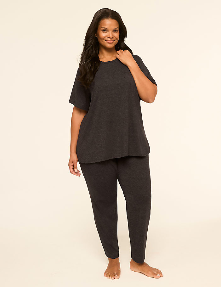 A smiling woman in a dark grey loungewear set stands against a plain, light-colored background.