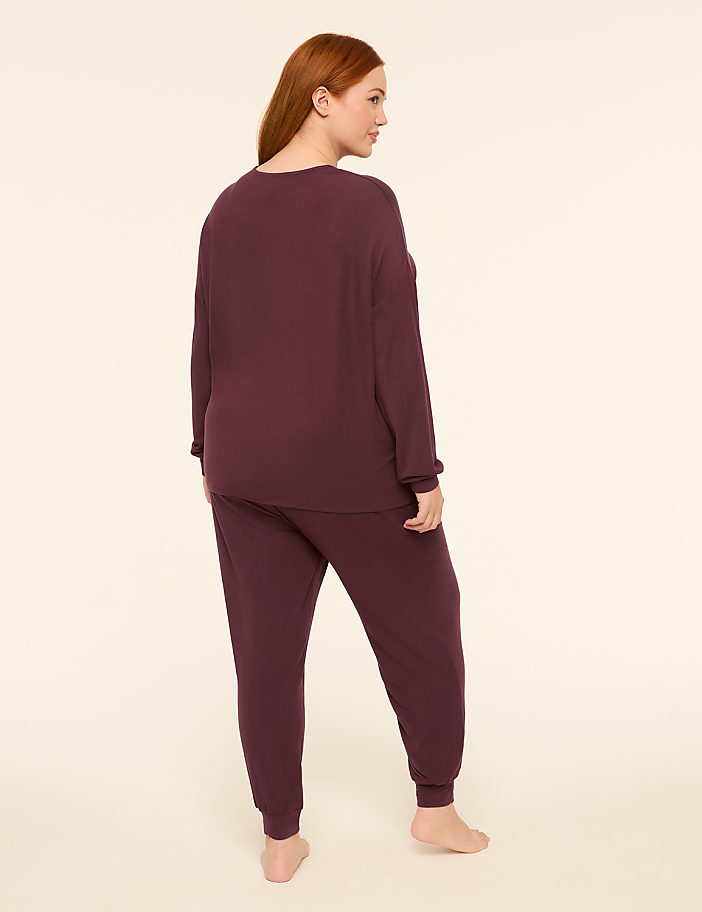 A woman with red hair stands facing away from the camera in a plum-colored loungewear set against a plain, light-colored background.